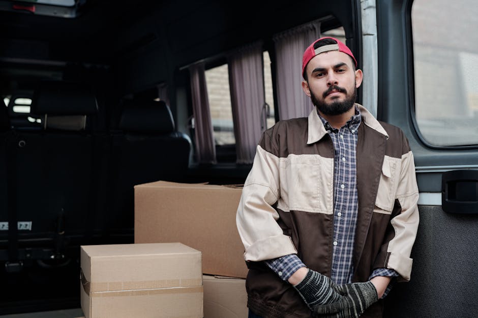 A man standing next to a partially loaded van during a home relocation, with several cardboard boxes of varying sizes arranged on the vehicle's interior floor. The man, dressed in a casual checkered shirt, a two-tone beige and brown jacket, and a red cap worn backwards, has a neutral expression and wears grey gloves, suggesting he is involved in the furniture transport and packing process. The van's sliding door is open, revealing internal space with curtains on the windows, and the exterior scene hints at a residential area. This image depicts professional removal services by Man With a Van Colindale, emphasizing the loading process and packing materials used in house removals.