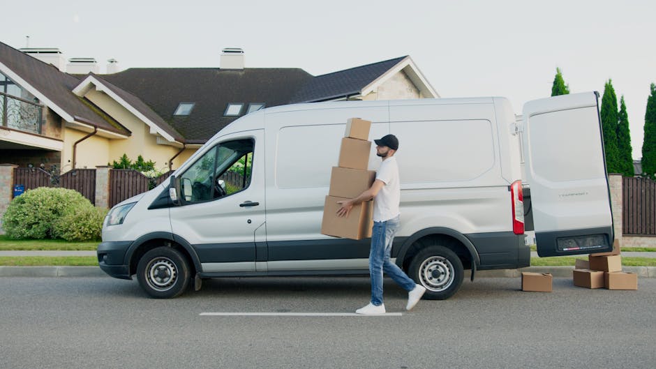 A man standing next to a partially loaded van during a home relocation, with several cardboard boxes of varying sizes arranged on the vehicle's interior floor. The man, dressed in a casual checkered shirt, a two-tone beige and brown jacket, and a red cap worn backwards, has a neutral expression and wears grey gloves, suggesting he is involved in the furniture transport and packing process. The van's sliding door is open, revealing internal space with curtains on the windows, and the exterior scene hints at a residential area. This image depicts professional removal services by Man With a Van Colindale, emphasizing the loading process and packing materials used in house removals.