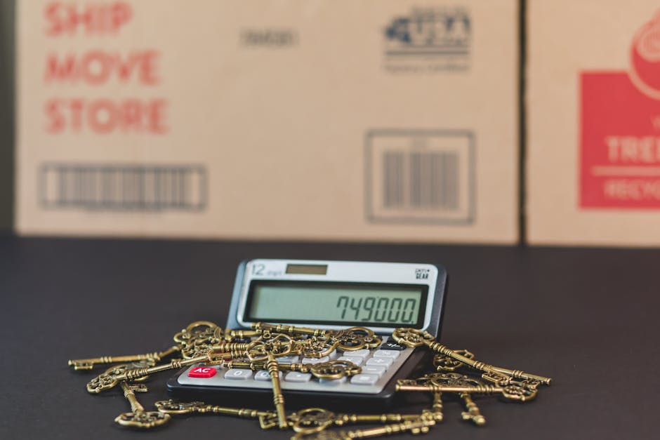 A calculator displaying the number 749800 is positioned on a black surface, with several old-fashioned brass keys scattered on top of it. Behind the calculator, there are multiple cardboard boxes with printed labels, including one with the words 'Ship', 'Move', and 'Store' in red text, and barcode labels. The scene appears to be set indoors, with the boxes arranged in the background, suggesting the process of packing for a home relocation. The brass keys, which resemble antique or skeleton keys, are casually placed, indicating a period of packing or decluttering. This image reflects aspects of furniture transport and packing during a professional house removal, as led by [COMPANY_NAME], with logistics involving boxes, packaging materials, and initial loading preparations. The lighting is neutral, and the overall setting emphasizes moving preparations and the logistical steps involved in house removals and furniture transport services.