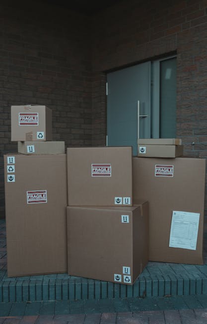 A calculator displaying the number 749800 is positioned on a black surface, with several old-fashioned brass keys scattered on top of it. Behind the calculator, there are multiple cardboard boxes with printed labels, including one with the words 'Ship', 'Move', and 'Store' in red text, and barcode labels. The scene appears to be set indoors, with the boxes arranged in the background, suggesting the process of packing for a home relocation. The brass keys, which resemble antique or skeleton keys, are casually placed, indicating a period of packing or decluttering. This image reflects aspects of furniture transport and packing during a professional house removal, as led by [COMPANY_NAME], with logistics involving boxes, packaging materials, and initial loading preparations. The lighting is neutral, and the overall setting emphasizes moving preparations and the logistical steps involved in house removals and furniture transport services.