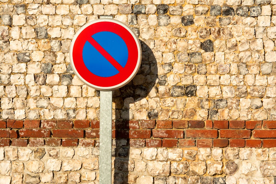 Close-up view of a round no parking traffic sign with a red border and blue centre, featuring a red diagonal line across it, mounted on a grey metal pole attached to a textured brick wall made of beige and brown stones. The sign casts a distinct shadow onto the wall, indicating strong sunlight. The brick wall has a horizontal reddish-brown brick line near the bottom, contrasting with the lighter stone surface above. The image captures a street scene related to urban regulations, which may be relevant in the context of house removals or moving services provided by Man With a Van Colindale, especially when considering parking restrictions during the landscaping or furniture transport phases of home relocation.