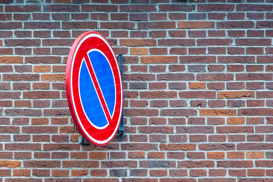 A round no parking sign mounted on a metal bracket attached to a brick wall, featuring a red border, a blue background, and a red diagonal line across the center. The brick wall behind it is composed of reddish-brown bricks with light gray mortar, typical of residential or commercial building exteriors. The sign is positioned near a doorway or wall surface, with no other objects visible in the image. This type of signage is relevant in the context of home relocation or furniture transport, indicating parking restrictions that may affect loading and unloading during house removals, and is associated with the services provided by Man With a Van Colindale, especially in relation to adhering to local council regulations such as those outlined for Bulky Item Rules in Colindale.