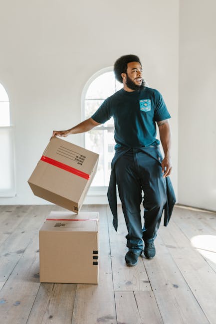 A man with dark curly hair and a beard, dressed in a navy blue T-shirt and dark work pants with a jumper tied around his waist, is inside a bright, spacious room with white walls and a large arched wi
