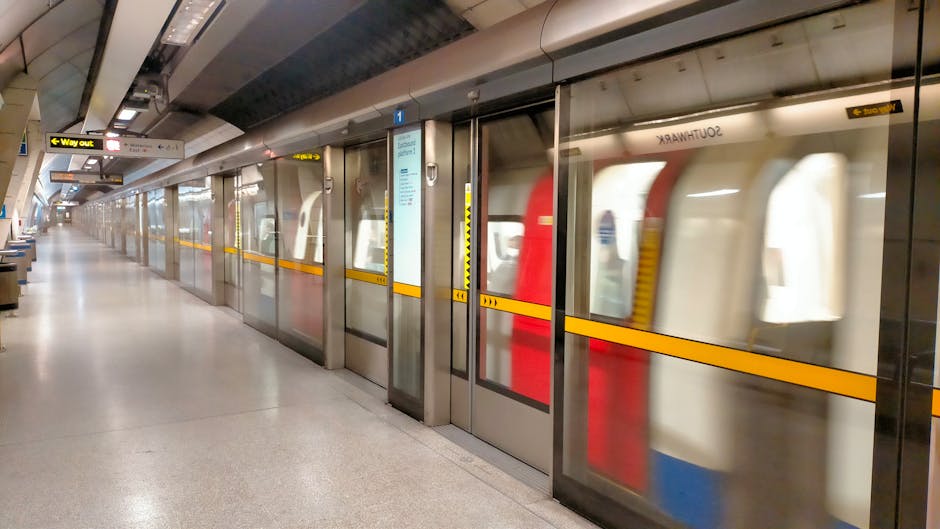 A long, well-lit underground station platform with a row of glass doors separating the waiting area from the train track. Behind the doors, a train with white and red exterior panels is visible, slightly blurred due to motion. The platform has a smooth, light-colored floor and several seating benches along its edge. Overhead, there are signs indicating directions and exits, including a yellow 'Way Out' sign. The station appears clean and modern, with metallic and glass infrastructural elements. This setting reflects the typical environment encountered during home relocation or moving processes, especially when coordinating transport to or from a tube station as part of a house removals service. Man With a Van Colindale is involved in logistical planning for local removals and moving projects, which may include arrangements for accessing transportation hubs like the Colindale Tube Station.