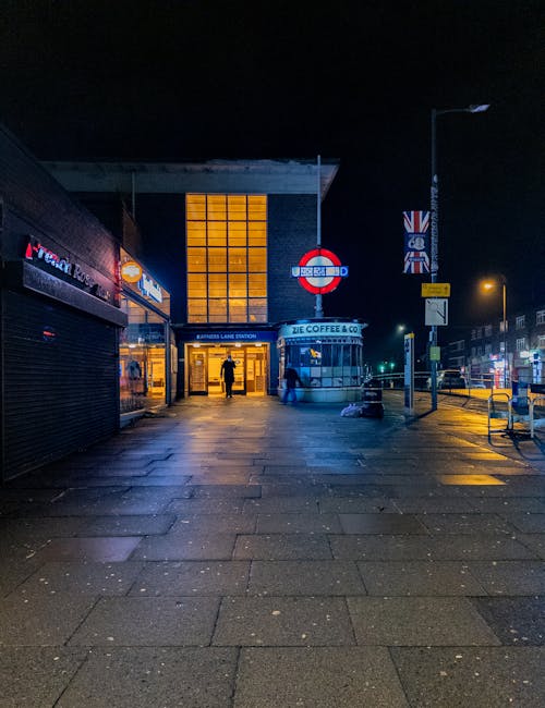 A nighttime view of the entrance to Colindale Tube Station showing illuminated signage and the station's glass facade with a glowing yellow-orange interior. In front of the station, the paved pavement is wet, reflecting nearby lights. To the right, a street lamp and various street signs, including one with the UK flag, are visible. The street is quiet with a few pedestrians near the station entrance, and the surrounding buildings are brick structures with shop fronts. This scene depicts an urban setting suitable for local home relocations or furniture transport, and the station's proximity indicates its importance in the moving logistics process, as handled by companies like Man With a Van Colindale.