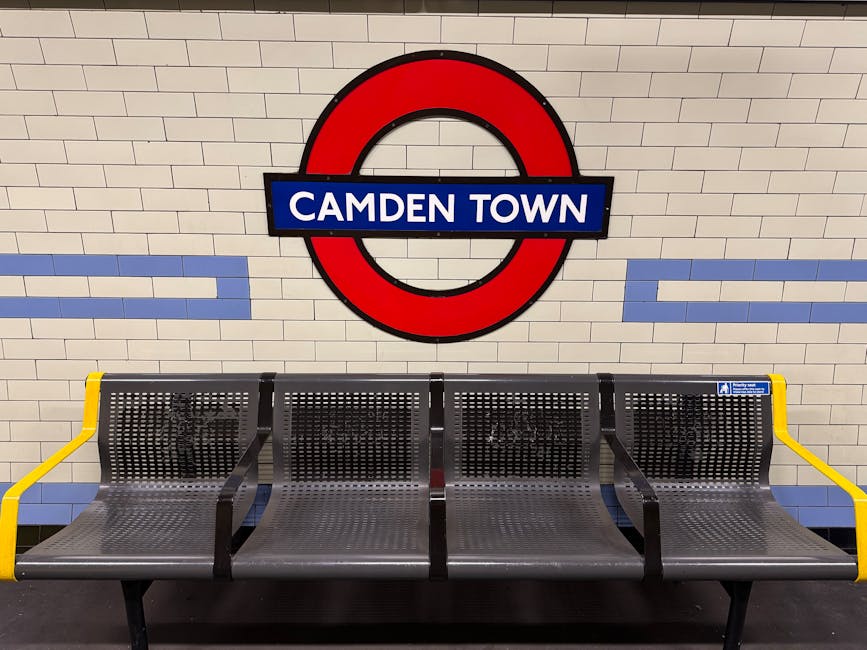 A long, well-lit underground station platform with a row of glass doors separating the waiting area from the train track. Behind the doors, a train with white and red exterior panels is visible, slightly blurred due to motion. The platform has a smooth, light-colored floor and several seating benches along its edge. Overhead, there are signs indicating directions and exits, including a yellow 'Way Out' sign. The station appears clean and modern, with metallic and glass infrastructural elements. This setting reflects the typical environment encountered during home relocation or moving processes, especially when coordinating transport to or from a tube station as part of a house removals service. Man With a Van Colindale is involved in logistical planning for local removals and moving projects, which may include arrangements for accessing transportation hubs like the Colindale Tube Station.