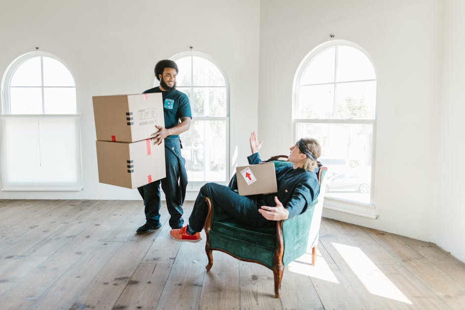 A professional removal team from Man With a Van Colindale is engaged in a home relocation process inside a bright, spacious room with white walls and large, arched windows allowing natural light to fill the space. One mover, a young man with curly hair and a beard, is standing indoors, carrying two large cardboard boxes secured with red and black tape, with an expression of focus. The other team member, an older woman with glasses and blonde hair, is seated comfortably in an upholstered armchair and holding a medium-sized cardboard box with a white arrow label on it, raising her hand in a friendly gesture. The room features a wooden floor and minimal furnishings, emphasizing the furniture and packing materials involved in the packing and moving process. Visible elements include the boxes, packing tape, and the way the items are being carefully loaded for transport, indicative of a furniture transport and packing operation by Man With a Van Colindale as part of a comprehensive house removal service.