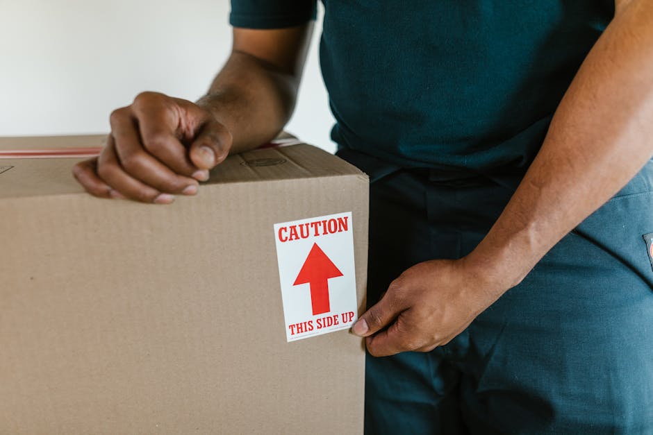 Close-up of a person wearing a dark green shirt and blue work trousers, securing a cardboard box with a red and white caution label that reads 'CAUTION THIS SIDE UP' with an upward arrow, indicating proper handling during a home relocation. The individual is holding the box with one hand while the other hand stabilizes it, positioned near a doorway or inside a property. In the background, part of a beige or light-colored wall is visible, suggesting an indoor environment. The scene reflects the careful packing and preparing process involved in furniture transport and moving services, such as those provided by Man With a Van Colindale, which specializes in house removals and packing during a professional move.