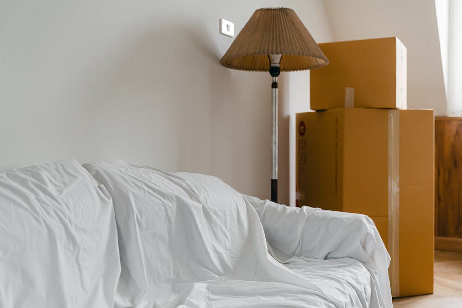 A professional removal team from Man With a Van Colindale is engaged in a home relocation process inside a bright, spacious room with white walls and large, arched windows allowing natural light to fill the space. One mover, a young man with curly hair and a beard, is standing indoors, carrying two large cardboard boxes secured with red and black tape, with an expression of focus. The other team member, an older woman with glasses and blonde hair, is seated comfortably in an upholstered armchair and holding a medium-sized cardboard box with a white arrow label on it, raising her hand in a friendly gesture. The room features a wooden floor and minimal furnishings, emphasizing the furniture and packing materials involved in the packing and moving process. Visible elements include the boxes, packing tape, and the way the items are being carefully loaded for transport, indicative of a furniture transport and packing operation by Man With a Van Colindale as part of a comprehensive house removal service.