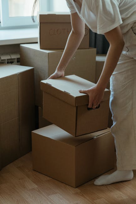 A person wearing a white shirt and light-colored trousers is in the process of organizing or packing moving boxes inside a home. The individual is holding a medium-sized cardboard box while standing on a wooden floor near a window with natural daylight illuminating the space. Several other cardboard boxes of various sizes are stacked around, with one labeled 'CLOTH' visible on the top. The boxes are made of plain brown cardboard, some with flaps closed and others open, suggesting packing or unpacking activities typical of a home relocation. Equipment such as a trolley or moving straps are not visible, but the setting appears tidy and prepared for the moving process. The background features a wall and window, indicating this activity takes place inside a residential interior as part of furniture transport and packing efforts coordinated by Man With a Van Colindale, supporting efficient house removals and relocation services.