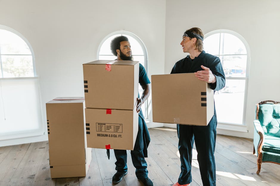 A person wearing a white shirt and light-colored trousers is in the process of organizing or packing moving boxes inside a home. The individual is holding a medium-sized cardboard box while standing on a wooden floor near a window with natural daylight illuminating the space. Several other cardboard boxes of various sizes are stacked around, with one labeled 'CLOTH' visible on the top. The boxes are made of plain brown cardboard, some with flaps closed and others open, suggesting packing or unpacking activities typical of a home relocation. Equipment such as a trolley or moving straps are not visible, but the setting appears tidy and prepared for the moving process. The background features a wall and window, indicating this activity takes place inside a residential interior as part of furniture transport and packing efforts coordinated by Man With a Van Colindale, supporting efficient house removals and relocation services.