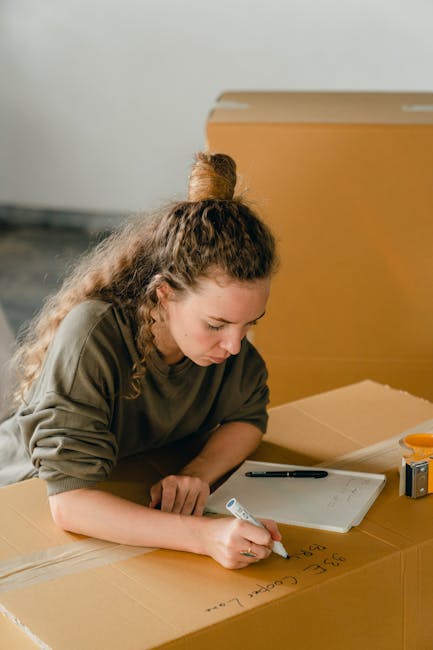 A woman with long, curly brown hair tied up in a bun is inside a room, leaning over a large cardboard box on a wooden surface. She is using a black marker to label the box, which already has some handwritten notes on it. The room has neutral walls, and in the background, there is a partially visible cardboard box and an orange object, possibly a tape dispenser or small toolbox. This scene illustrates the packing and moving process, typical of home relocation services provided by Man With a Van Colindale, with the woman preparing boxes for transport. The overall setting emphasizes careful packing, labeling, and the planning involved in furniture transport and household packing during house removals, supporting the context of estimating moving costs and logistics in the NW9 postcode area.