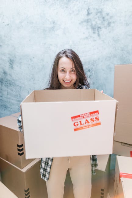 A woman with long brown hair, smiling and looking directly at the camera, stands indoors against a plain, textured light blue wall. She is holding an open, empty cardboard box labeled 'GLASS' in red print, which is positioned at chest height. Around her, there are several other closed and open cardboard boxes of various sizes, some with black tape or red labels, arranged on the floor and nearby surfaces. The scene depicts a home relocation or packing and moving process, likely involving a professional removals service such as Man With a Van Colindale. The lighting is bright and natural, emphasizing the orderly loading environment as part of furniture transport and move logistics.
