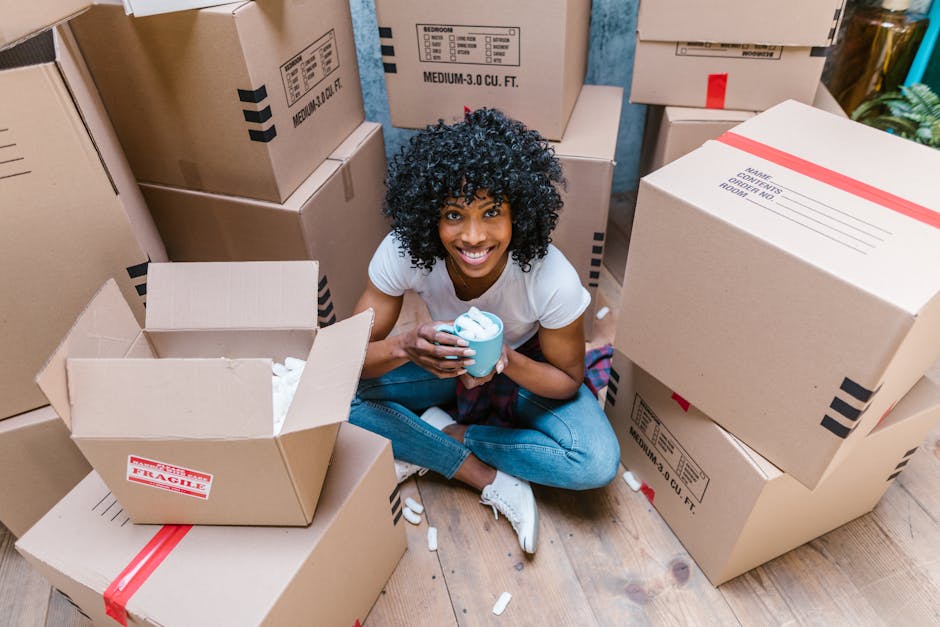A woman with long, curly brown hair tied up in a bun is inside a room, leaning over a large cardboard box on a wooden surface. She is using a black marker to label the box, which already has some handwritten notes on it. The room has neutral walls, and in the background, there is a partially visible cardboard box and an orange object, possibly a tape dispenser or small toolbox. This scene illustrates the packing and moving process, typical of home relocation services provided by Man With a Van Colindale, with the woman preparing boxes for transport. The overall setting emphasizes careful packing, labeling, and the planning involved in furniture transport and household packing during house removals, supporting the context of estimating moving costs and logistics in the NW9 postcode area.