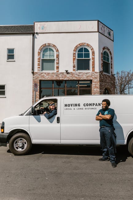 A white moving van with the branding 'MOVING COMPANY LOCAL & LONG DISTANCE' parked in front of a modern, multi-story residential building during a home relocation process. Inside the driver’s cab, a person wearing sunglasses and a cap is visible, possibly communicating with the second worker standing beside the vehicle. The second worker, dressed in a blue shirt and dark trousers, is standing with arms crossed next to the van, engaged in conversation. The building features a combination of white plaster and brickwork, with large windows and slight decorative arches above the windows on the upper floor. The scene is outdoors on a clear day, with the pavement and part of the building's entrance visible, indicating the loading or unloading phase of a house move. This image exemplifies furniture transport, packing, and loading activities typical of house removals services provided by companies such as Man With a Van Colindale, supporting detailed and accessible content on relocation logistics.