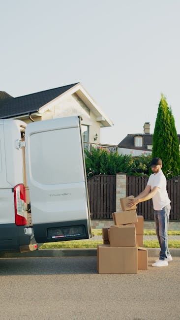 A man wearing a white t-shirt and dark trousers is seen standing next to an open white moving van parked on a residential street. He is handling cardboard boxes, some stacked on the pavement and others being loaded or unloaded from the vehicle. The van's rear doors are fully open, revealing the interior space used for furniture and household items during a house move. In the background, there is a modern house with a gabled roof, surrounded by greenery including a tall coniferous tree and a wooden fence. The scene takes place during daylight with clear weather, capturing a typical home relocation process supported by Man With a Van Colindale, emphasizing packing and furniture transport for house removals.