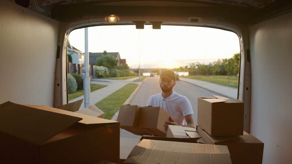 A white moving van with the branding 'MOVING COMPANY LOCAL & LONG DISTANCE' parked in front of a modern, multi-story residential building during a home relocation process. Inside the driver’s cab, a person wearing sunglasses and a cap is visible, possibly communicating with the second worker standing beside the vehicle. The second worker, dressed in a blue shirt and dark trousers, is standing with arms crossed next to the van, engaged in conversation. The building features a combination of white plaster and brickwork, with large windows and slight decorative arches above the windows on the upper floor. The scene is outdoors on a clear day, with the pavement and part of the building's entrance visible, indicating the loading or unloading phase of a house move. This image exemplifies furniture transport, packing, and loading activities typical of house removals services provided by companies such as Man With a Van Colindale, supporting detailed and accessible content on relocation logistics.