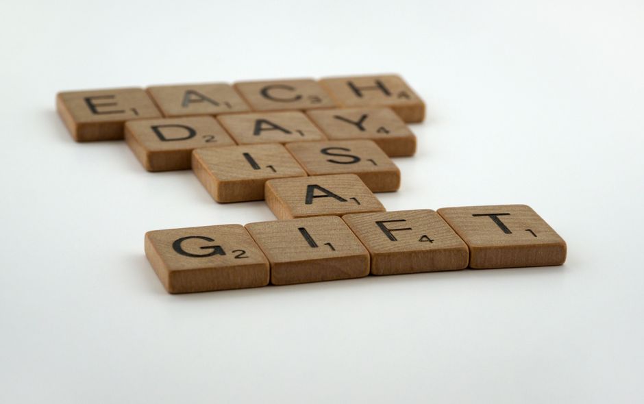 A close-up image displays wooden Scrabble tiles spelling out the words 'EACH DAY' and 'GIFT' on a white surface. The tiles are arranged in two lines, with 'EACH DAY' placed above and slightly to the left of 'GIFT'. The background is plain and well-lit, emphasizing the natural wood texture of the tiles and the black printed letters. This image relates to themes of daily routines or special moments, which can be relevant in the context of house removals and home relocation services offered by Man With a Van Colindale, especially when discussing moving plans or packing logistics for same-day urgent removals in Colindale.