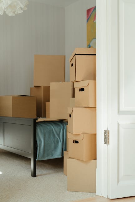 A room with several stacked cardboard moving boxes of various sizes placed atop a blue fabric covering a grey wooden sideboard. The boxes are arranged in a narrow corridor beside an open white door leading to an interior space. The walls are decorated with a colorful abstract painting, and the room is well-lit, likely during a home relocation process. The image captures the scene in a domestic setting, illustrating the packing and moving stage for a house removal service, with boxes waiting to be transported. This visual is relevant to the services offered by Man With a Van Colindale, emphasizing furniture transport and packing logistics during tight access moves in Colindale flats.