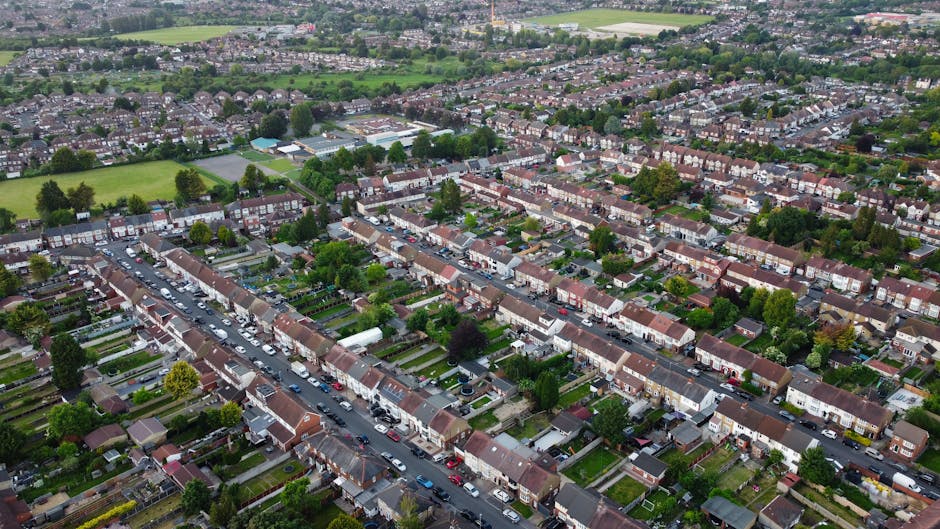 An aerial view of a residential neighborhood in Colindale showing rows of terraced houses with small front gardens, parked cars along the narrow streets, and a few larger green spaces and playing fields in the background. The image captures the densely packed housing area with uniform rooftops, surrounded by trees and greenery, under an overcast sky. This scene illustrates the typical layout of homes that may require precise planning for home relocation and furniture transport during tight access moves, with Man With a Van Colindale providing expert removals services to navigate such environments.