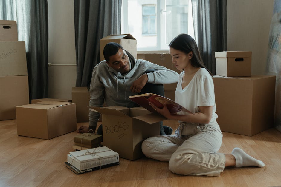 A room with several stacked cardboard moving boxes of various sizes placed atop a blue fabric covering a grey wooden sideboard. The boxes are arranged in a narrow corridor beside an open white door leading to an interior space. The walls are decorated with a colorful abstract painting, and the room is well-lit, likely during a home relocation process. The image captures the scene in a domestic setting, illustrating the packing and moving stage for a house removal service, with boxes waiting to be transported. This visual is relevant to the services offered by Man With a Van Colindale, emphasizing furniture transport and packing logistics during tight access moves in Colindale flats.